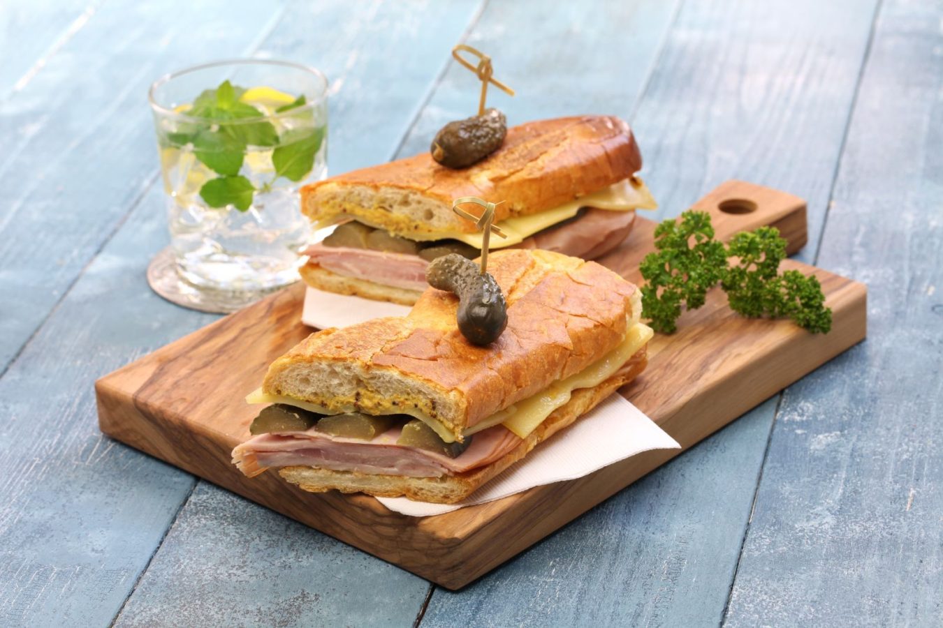 Cuban sandwich served with greens and pickles on a charcuterie board with a refreshing cocktail on the side